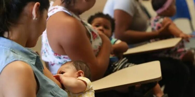Quatro mulheres amamentam seus bebês sentadas em carteiras escolares, ambiente de sala de aula