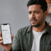 Homem de expressão preocupada mostra celular com saldo de R$500,00 em aplicativo bancário