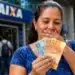 Mulher segura cédulas de real diante de uma agência da Caixa, representando a antecipação do Bolsa Família de julho.