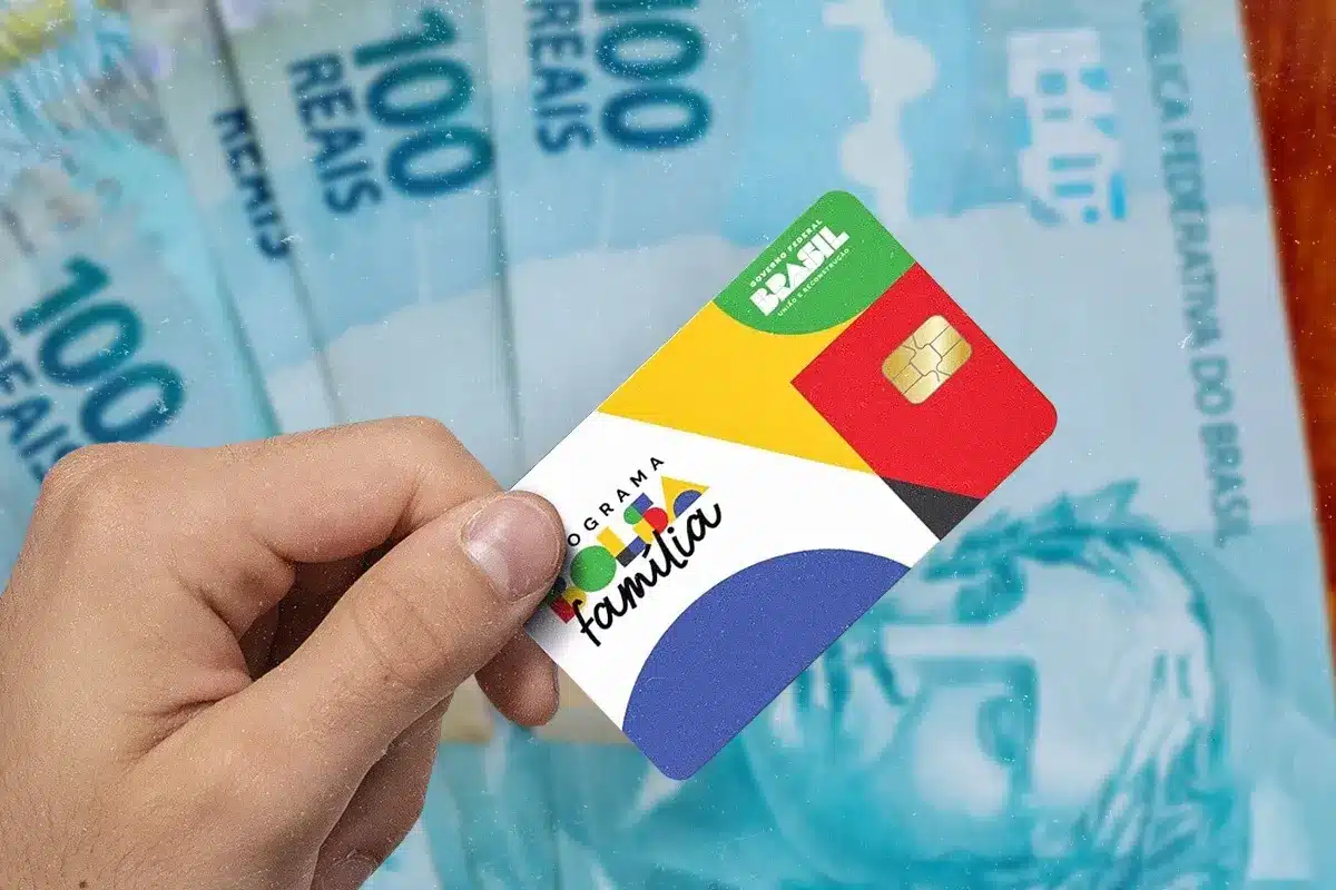 Mão segura cartão do Bolsa Família diante de cédulas de real, representando a movimentação antecipada do benefício.