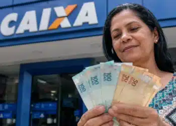 Mulher segura cédulas de real em frente a uma agência da Caixa, representando as novas liberações do Saque Calamidade do FGTS.