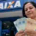 Saque Calamidade do FGTS ganha novas liberações; veja quem pode pedir R$ 6.220 a partir desta terça-feira (28) 14 Mulher segura cédulas de real em frente a uma agência da Caixa, representando as novas liberações do Saque Calamidade do FGTS.