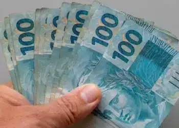 Mão segura cédulas de R$ 100, representando a solicitação do Saque Calamidade de até R$ 6.220, liberado pela Caixa.
