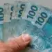 Mão segura cédulas de R$ 100, representando a solicitação do Saque Calamidade de até R$ 6.220, liberado pela Caixa.