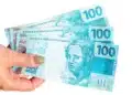 Mão segurando cédulas de real representando o pagamento de R$ 300 do programa Mães de Pernambuco
