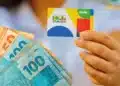 Beneficiária segura o cartão do Bolsa Família e notas de real, representando os pagamentos do benefício em agosto.