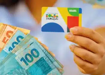 Beneficiária segura o cartão do Bolsa Família e notas de real, representando os pagamentos do benefício em agosto.