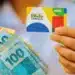 Bolsa Família de agosto será pago a partir de 18/8: Caixa define a ordem pelo final do NIS 14 Beneficiária segura o cartão do Bolsa Família e notas de real, representando os pagamentos do benefício em agosto.