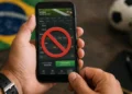 Celular com símbolo de bloqueio em plataforma de apostas representando restrição para beneficiários do Bolsa Família e BPC