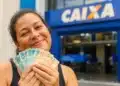 Trabalhadora segura cédulas de real em frente a uma agência da Caixa, representando o saque-aniversário do FGTS para nascidos em agosto.