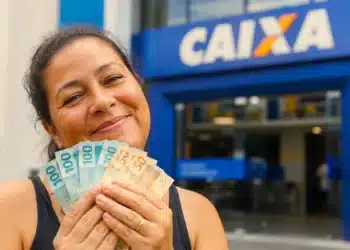 Trabalhadora segura cédulas de real em frente a uma agência da Caixa, representando o saque-aniversário do FGTS para nascidos em agosto.