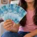 Filhos e dependentes de vítimas de feminicídio podem ter direito a pensão de R$ 1.621; veja como solicitar 14 Menina segurando notas de R$ 100, representando a pensão destinada a filhos e dependentes de vítimas de feminicídio.