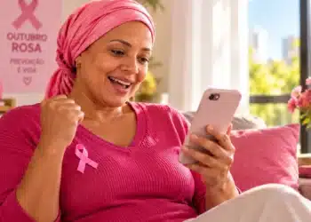 INSS concede benefícios por câncer de mama; saiba como solicitar pelo Meu INSS ou pelo telefone 6 Mulher com lenço rosa na cabeça e laço do Outubro Rosa na blusa sorri ao olhar para o celular, sentada no sofá de sua casa.