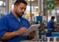 Fim da escala 6x1: veja como será feito o cálculo das horas de trabalho 14 Trabalhador de uniforme azul registra informações em uma prancheta dentro de uma fábrica, representando o controle das horas de trabalho com o fim da escala 6x1.