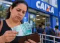 Saque Calamidade do FGTS: confira a lista de cidades habilitadas em outubro de 2026 e o prazo de cada uma 15 Mulher segura notas de R$ 100 e uma carteira em frente a uma agência da Caixa Econômica Federal, com pessoas aguardando atendimento ao fundo.
