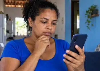 Beneficiária consulta o celular em casa com expressão pensativa, representando a consulta ao Bolsa Família de outubro.