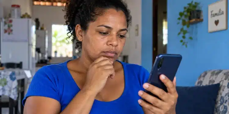 Beneficiária consulta o celular em casa com expressão pensativa, representando a consulta ao Bolsa Família de outubro.