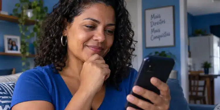 Mulher de blusa azul consulta informações pelo celular enquanto está sentada no sofá de casa.