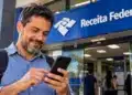 Receita libera consulta ao lote residual do IRPF 2026 nesta sexta (23); crédito sai no dia 30 14 Homem utilizando o celular em frente a uma unidade da Receita Federal, representando a consulta ao lote residual de restituição do IRPF 2026.