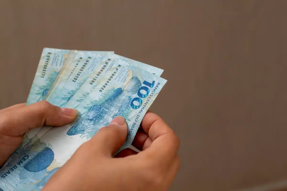 Décimo terceiro de 2026 sai mais cedo para quem tem carteira assinada; confira a data-limite 2 Mãos de uma pessoa contando cédulas de R$ 100, representando o recebimento do décimo terceiro salário e os descontos de INSS e Imposto de Renda.