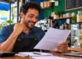 MEIs com dívidas inscritas na dívida ativa até 13 de outubro terão os débitos zerados a partir de janeiro 15 Microempreendedor individual sorri ao analisar documento financeiro em sua cafeteria, ilustrando a possibilidade de perdão de dívidas de MEIs.