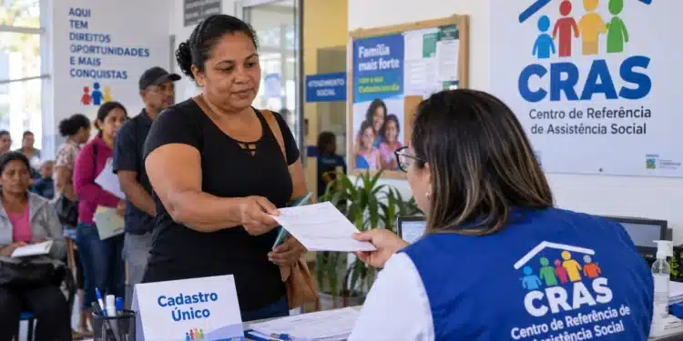 Mulher entrega documentos a uma atendente do CRAS durante atendimento do Cadastro Único, com moradores aguardando na fila.