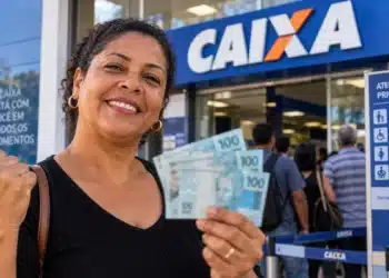 Mulher sorridente segura cédulas de R$ 100 em frente a uma agência da Caixa Econômica Federal, com clientes aguardando atendimento.