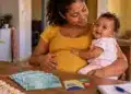Bolsa Família para nutriz e gestante: veja quantas parcelas de R$ 58 cada uma recebe e o valor total 2 Mulher gestante com bebê no colo diante de dinheiro e cartão do Bolsa Família sobre a mesa