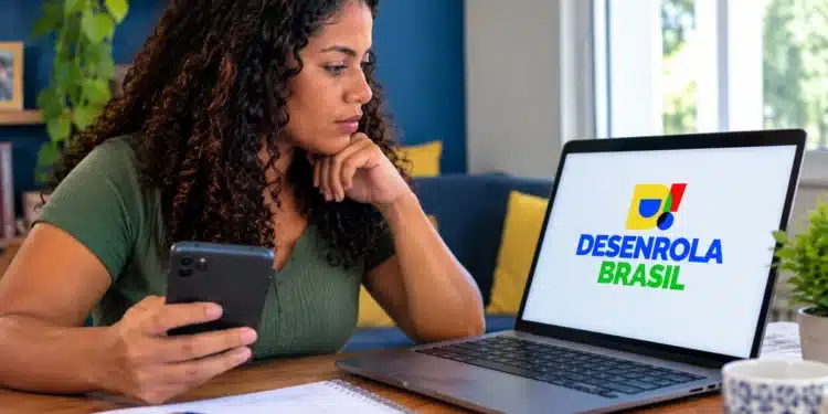 Mulher consulta o programa Desenrola Brasil no notebook enquanto segura o celular.