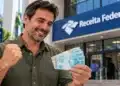 Homem segura cédulas de R$ 100 em frente à Receita Federal, ilustrando a restituição antecipada do Imposto de Renda.