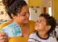 Mães de Pernambuco abre 2.760 vagas com auxílio de R$ 300 até 22 de outubro; veja quem pode se inscrever 14 Mãe segura cédulas de R$ 100 enquanto observa o filho em casa, ilustrando o auxílio de R$ 300 do programa Mães de Pernambuco.
